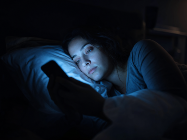 Person lying in dark bedroom scrolling phone, blue screen light glowing on face, illustrating blue light's effect on sleep